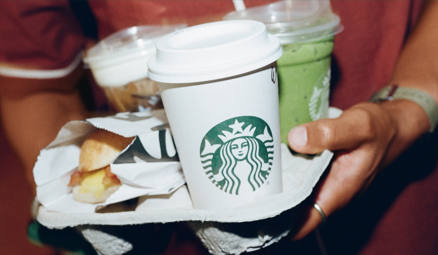 Order Starbucks® Delivery online with the Starbucks® app or website. A close-up of someone carrying a drink carrier full of Starbucks drinks and a hot breakfast sandwich.