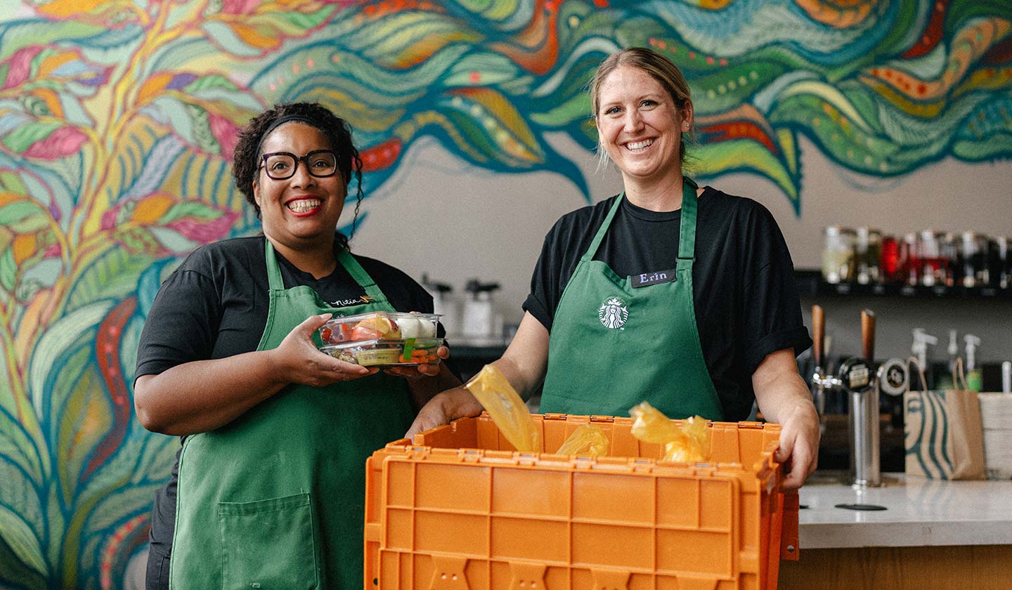 Two Starbucks employees in a Starbucks store smile for the camera while displaying a yellow crate of packaged food.