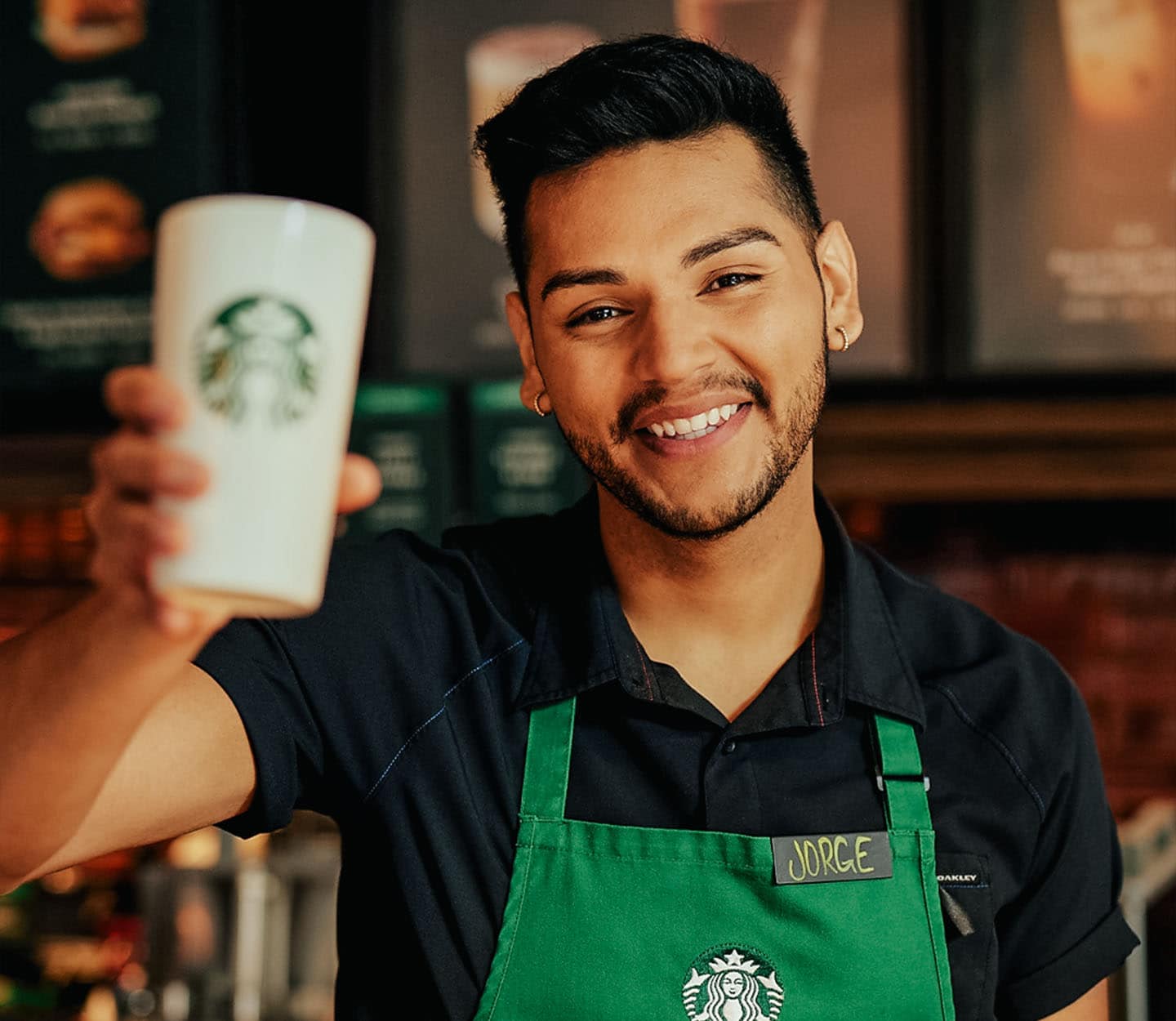 Starbucks barista in a green apron holds up a to-go cup while smiling at the camera.