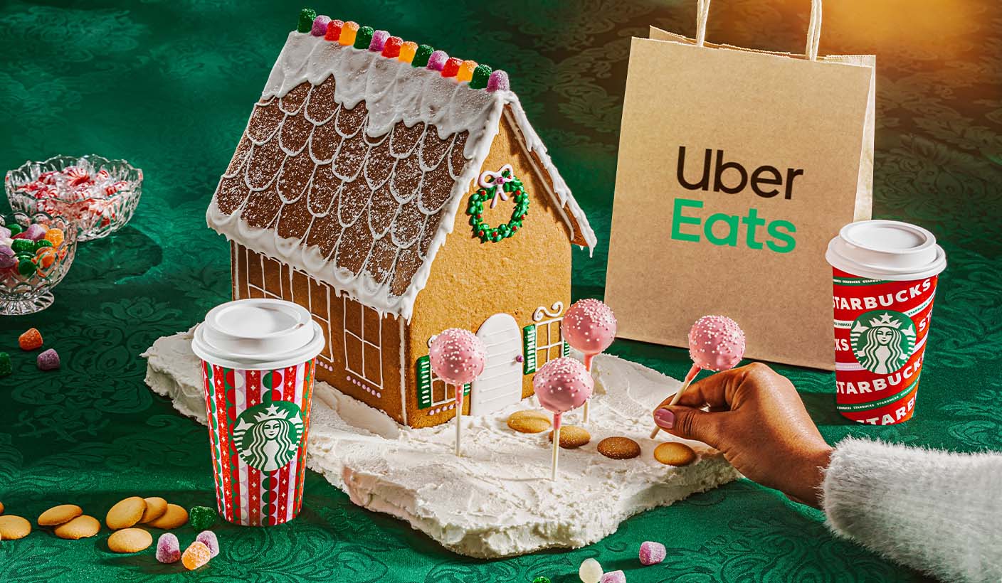 Holiday gingerbread house with Cake Pops whimsically serving as trees in house’s front yard. A hand places the Cake Pops while a Starbucks cup and Uber Eats bag stand on either side.
