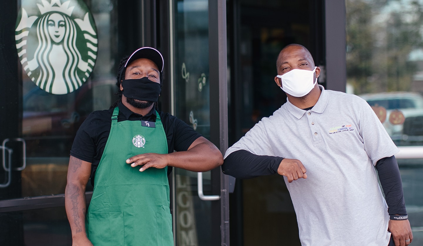 A Starbucks partner (employee) bumping elbows with a nonprofit business owner, both are wearing facial coverings standing outside of a Starbucks cafe and looking at camera.