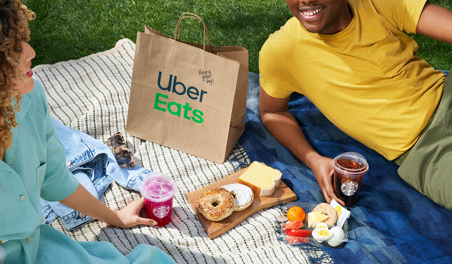 Two people featured on a picnic blanket with Starbucks products and an Uber Eats delivery bag.
