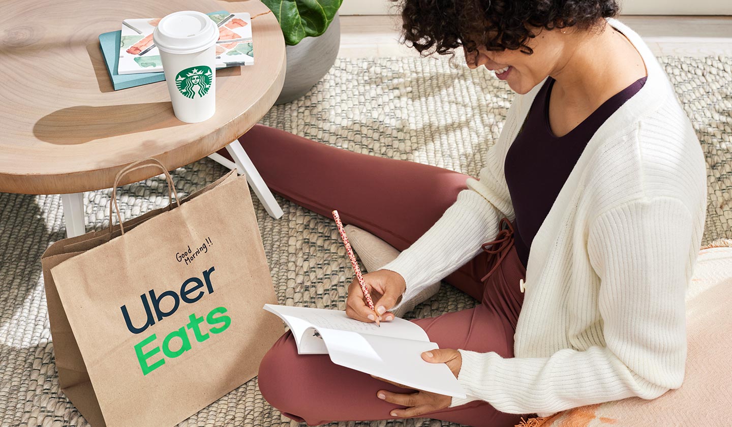 A woman sitting next to a coffee table writing in a journal. There is a Starbucks hot beverage sitting on top of the table, and an Uber Eats delivery bag on the floor.