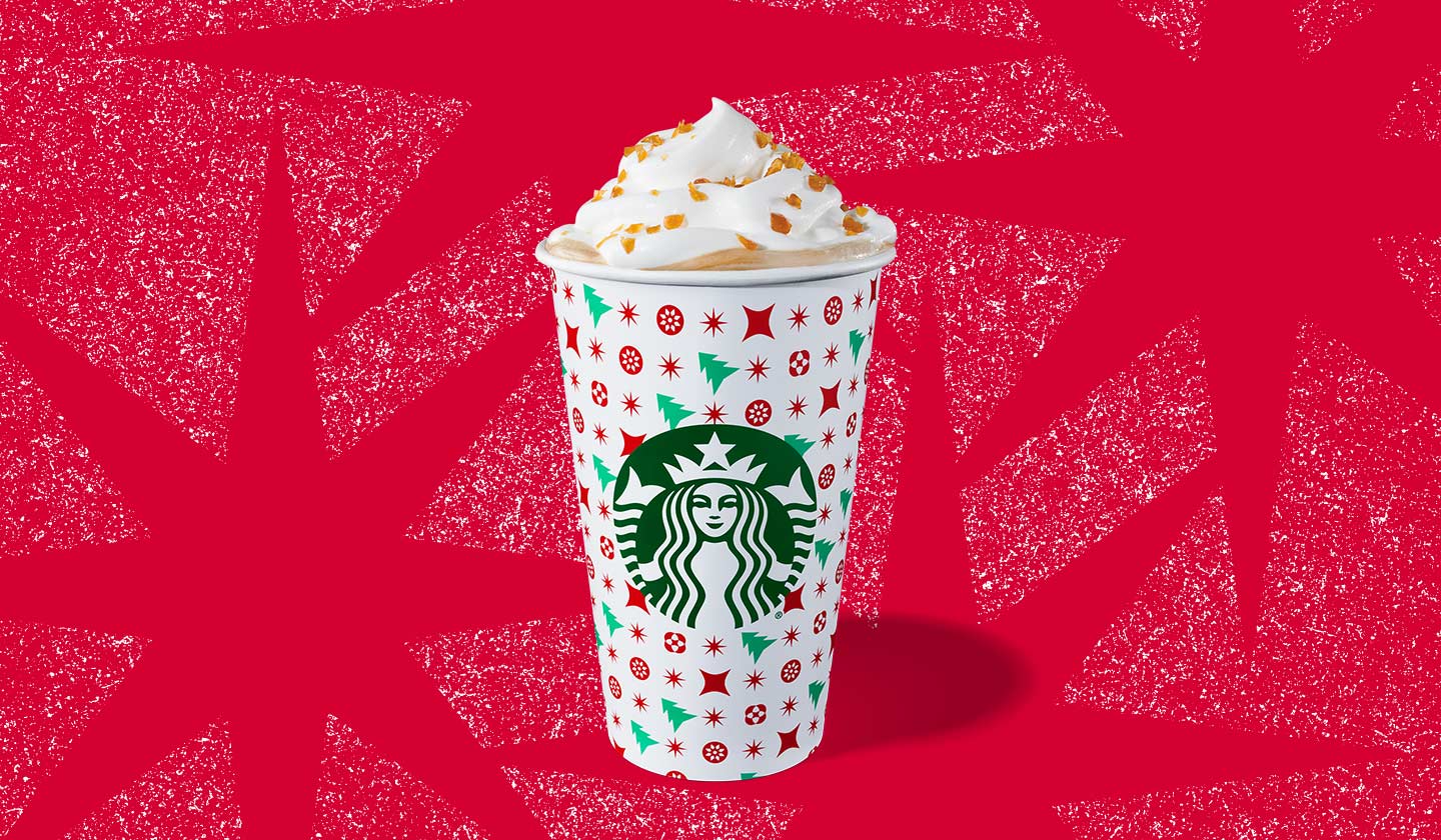 Hot espresso drink with whipped cream in a to-go cup with green trees and red stars on a white background.