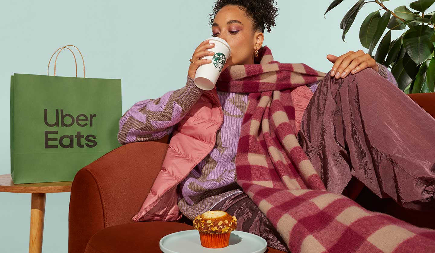 Person lounging on a couch wearing a comfortable scarf. The person is enjoying some Starbucks® delivered in a green Uber Eats bag is seen to the side.