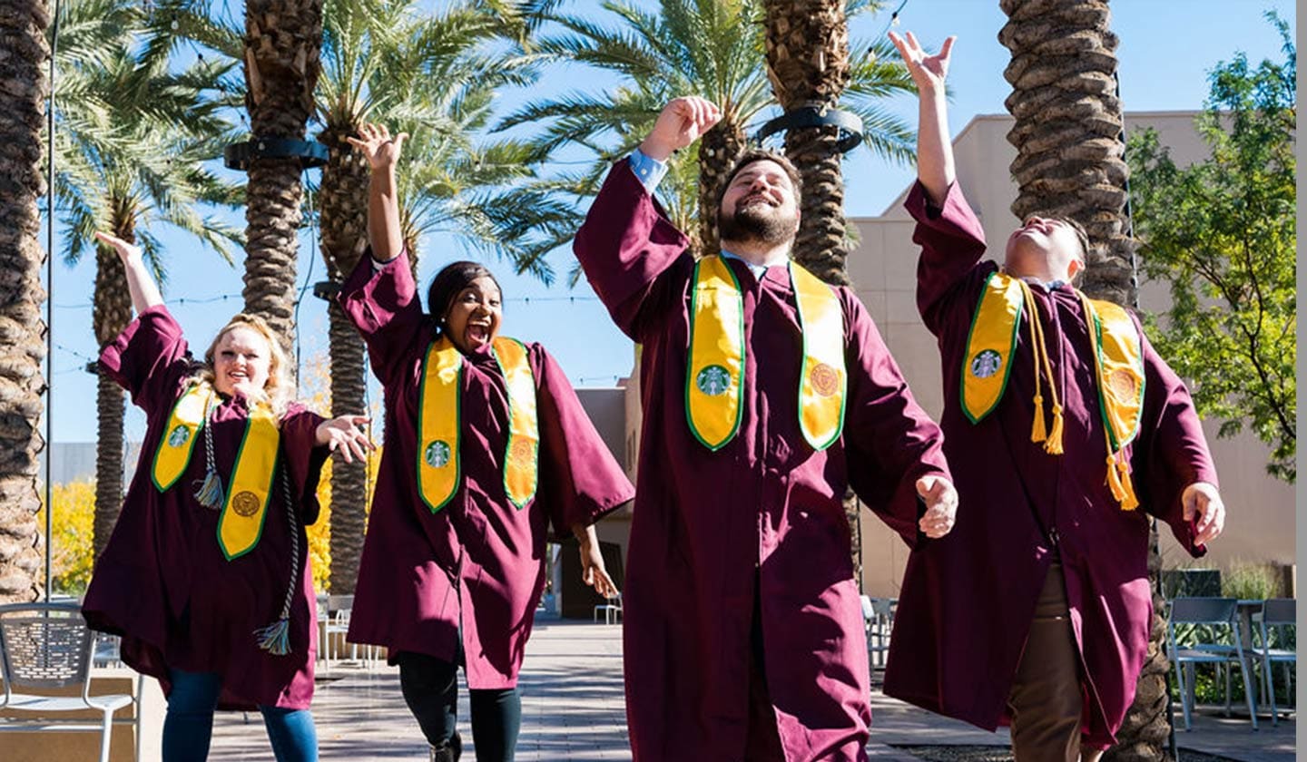 Four Starbucks College Achievement Plan graduates wearing maroon graduation robes and SCAP stoles throwing their caps in the air.
