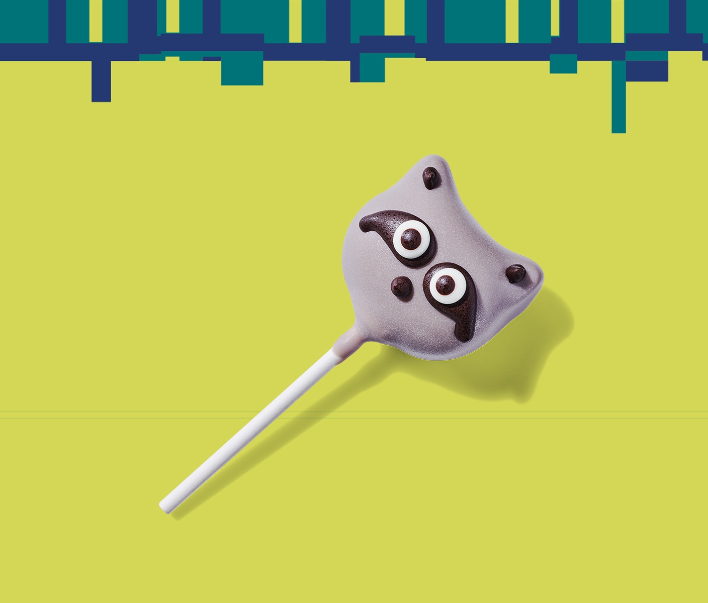 A cake pop on a stick in the shape of a raccoon head with big eyes atop a black band.