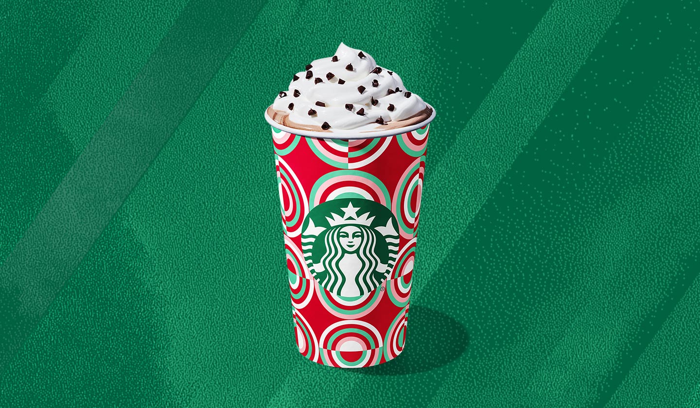 A hot coffee drink in a festive red and green patterned cup with a whipped cream topping and chocolate curls.