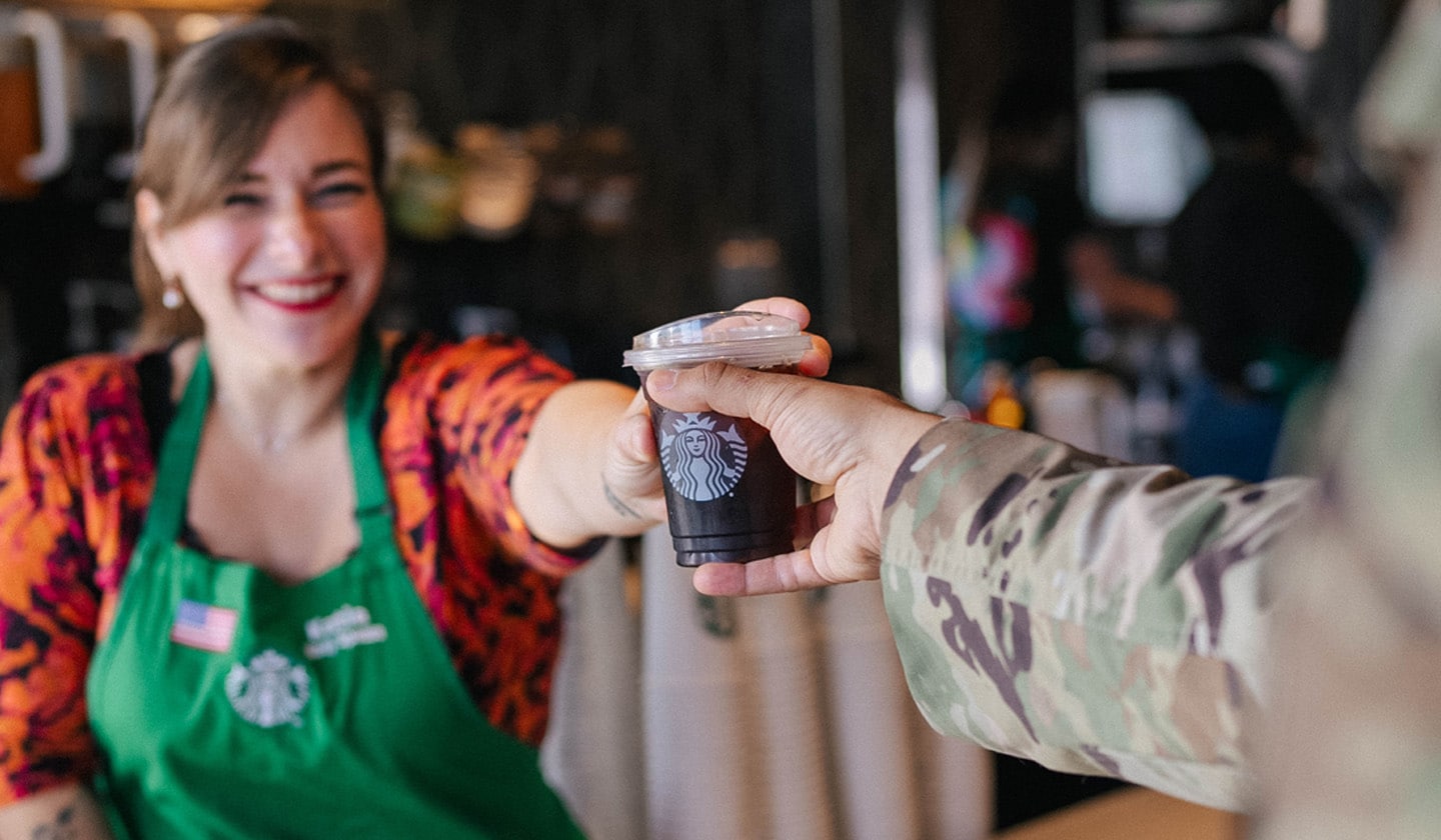 A smiling female Starbucks barista handing an iced coffee to a member of the military community.