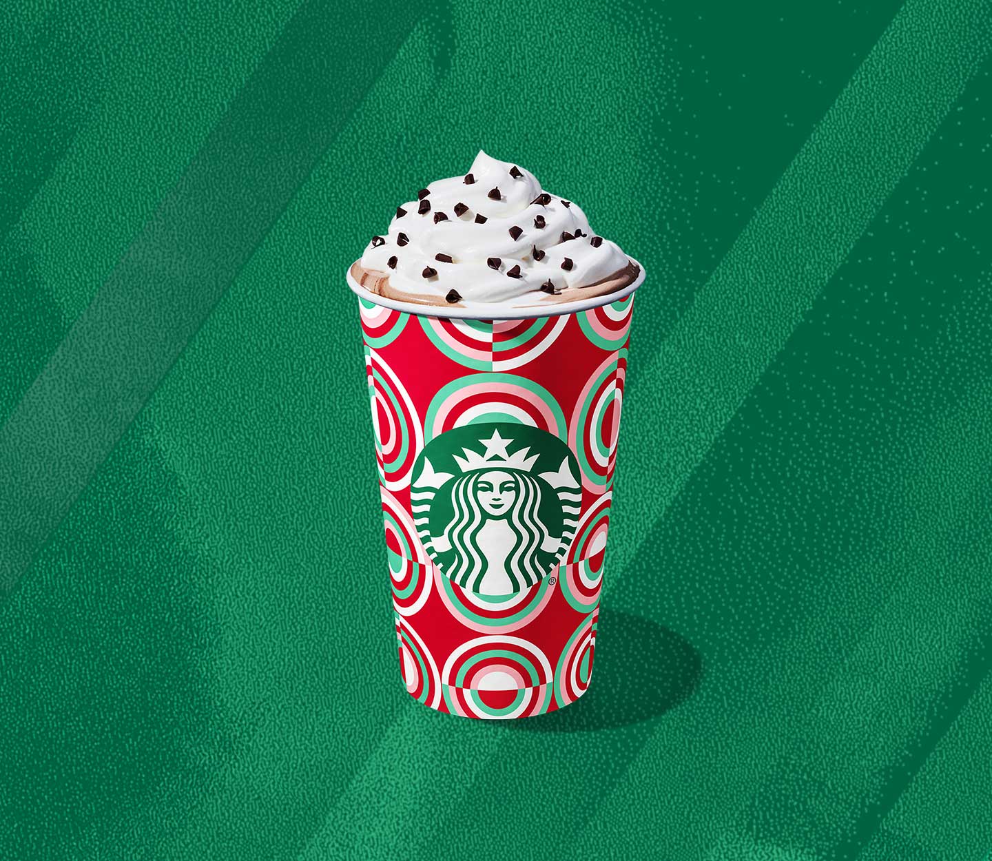 A hot coffee drink in a festive red and green patterned cup with whipped cream and chocolate curls on top.