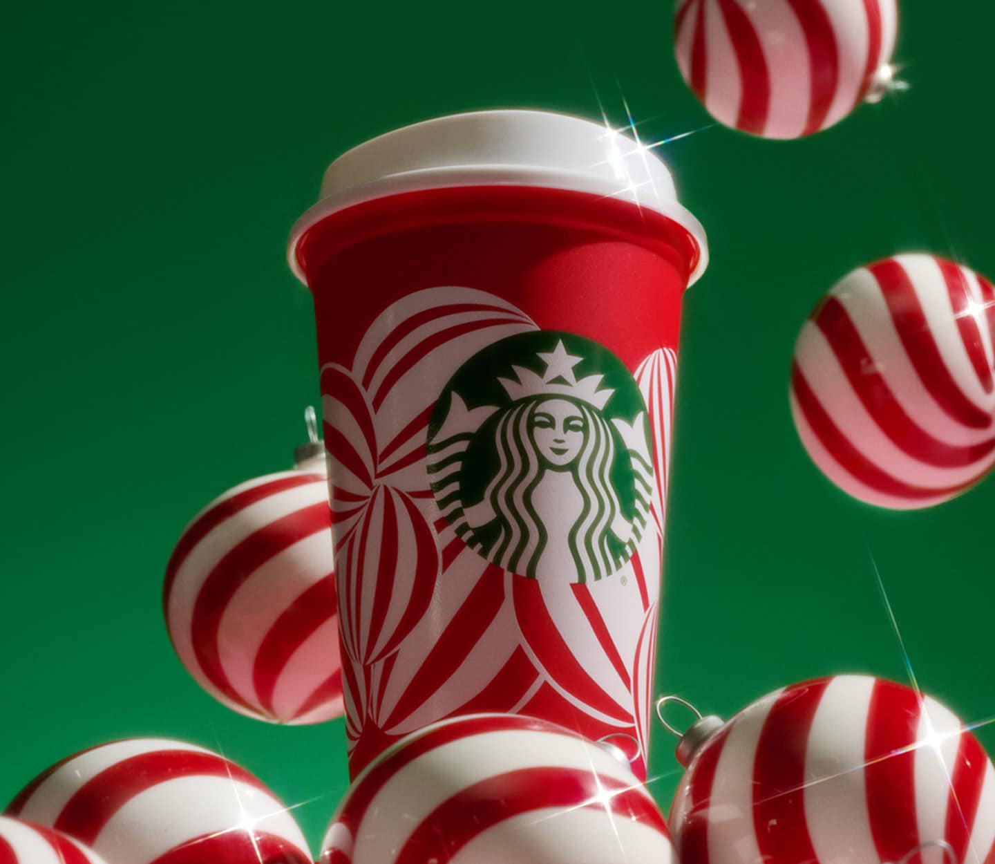 A reusable red cup with a red and white ornament design sits among a pile of red and white ornament balls, with more falling around it.