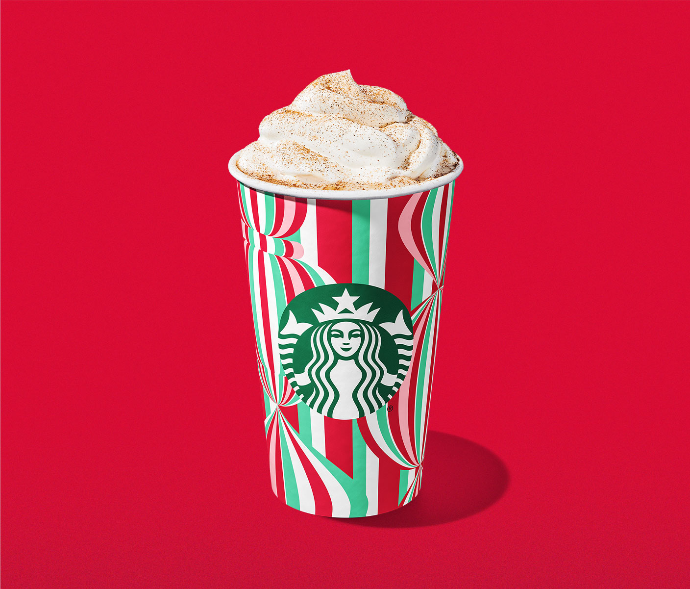 A hot coffee drink in a festive red and green patterned cup with whipped cream and pumpkin spice powder on top.