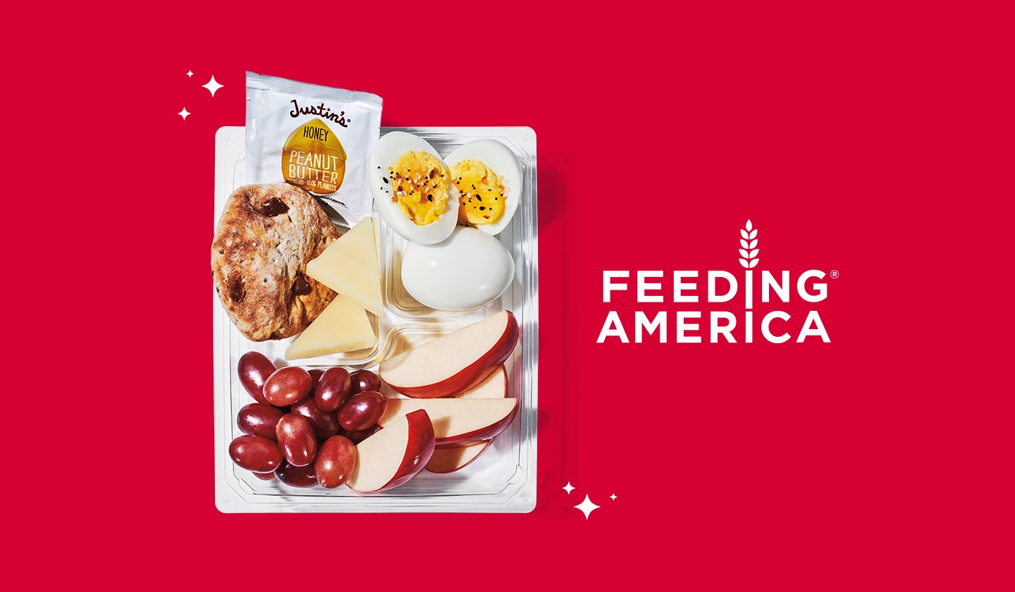 A Starbucks protein box on a red backdrop, next to the Feeding America logo. The protein box contains two hard-boiled eggs, apple slices, grapes, cheese slices, a piece of multigrain bread and a packet of honey peanut butter.