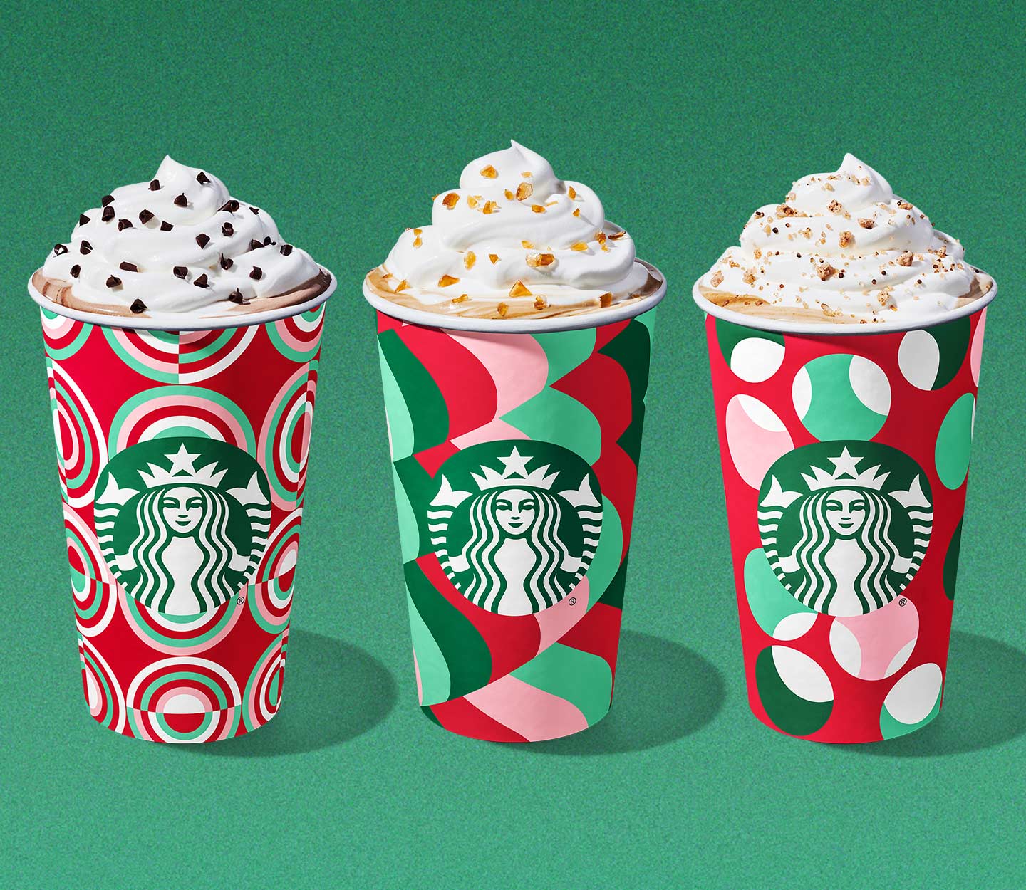 Three hot coffee drinks in festive red and green cups with whipped cream and holiday toppings sprinkled on top.