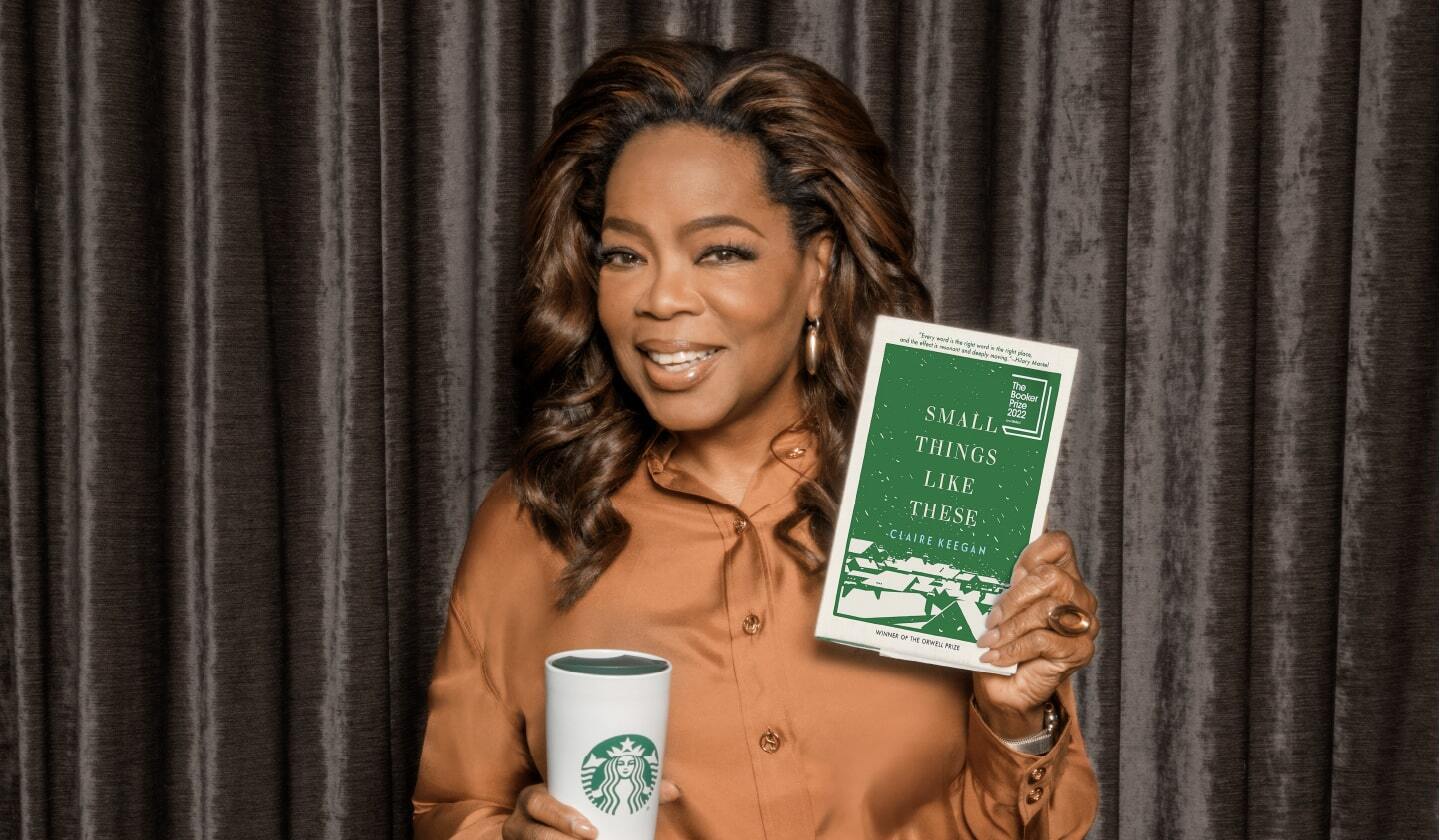 Oprah Winfrey holds a Starbucks coffee tumbler and shows the camera a book:  "Small Things Like These" by Claire Keegan.