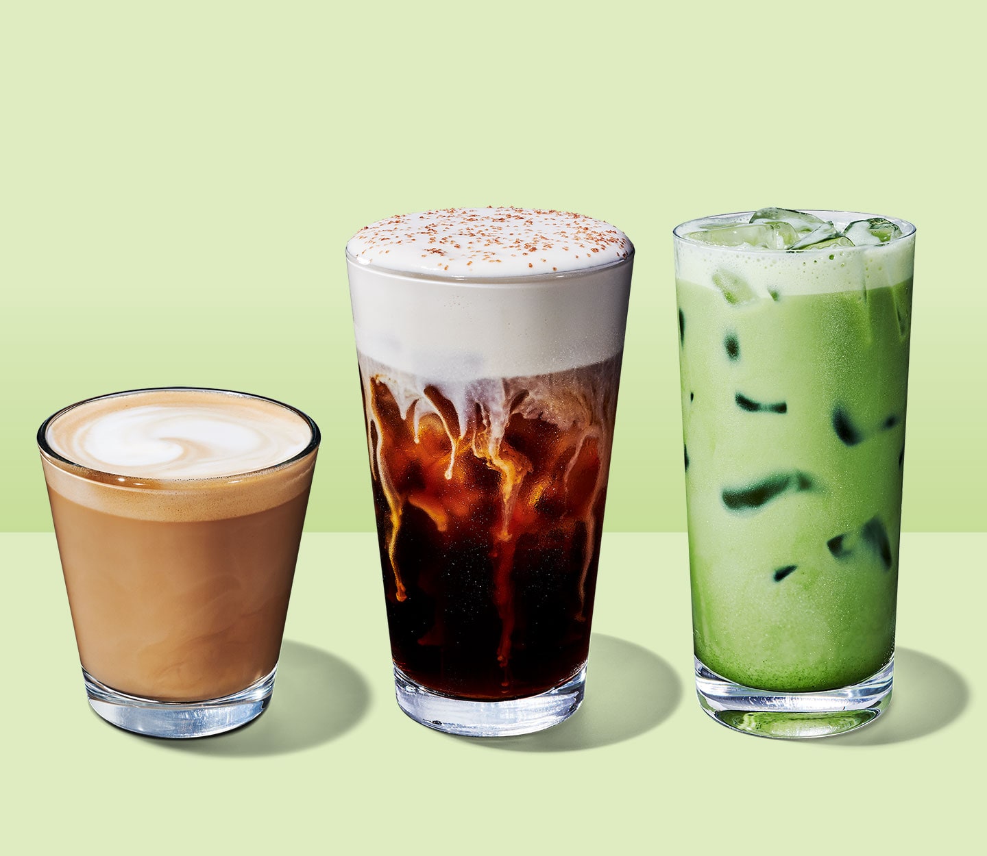 A trio of drinks in clear glasses: a short-sized creamy coffee drink with frothy top, a tall cold coffee drink with foam and sprinkles and a tall iced green tea drink.