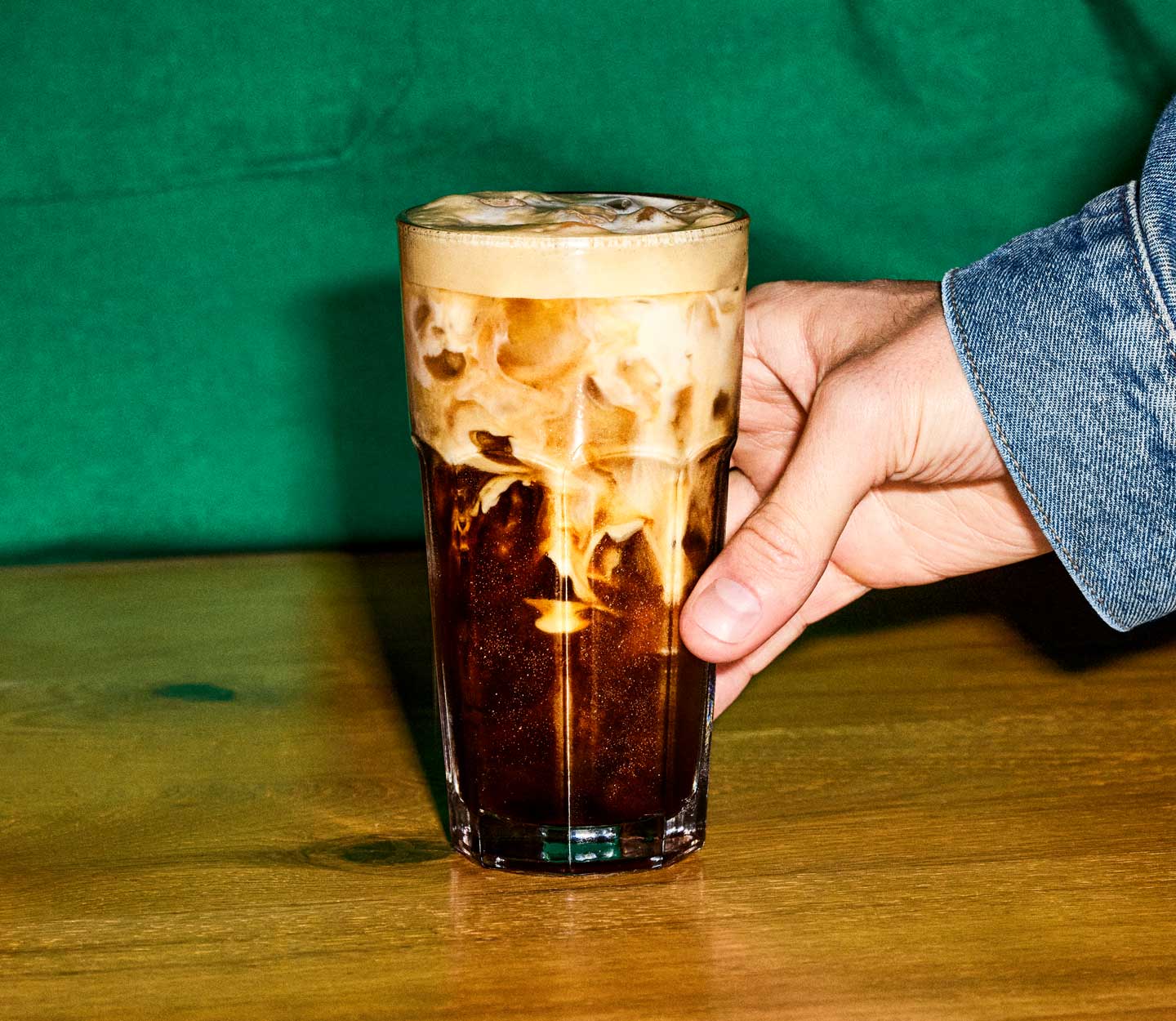 An icy coffee drink with creamy marbling on a counter is about to be picked up by a customer wearing a jean jacket. A barista in a green apron stands in the background.