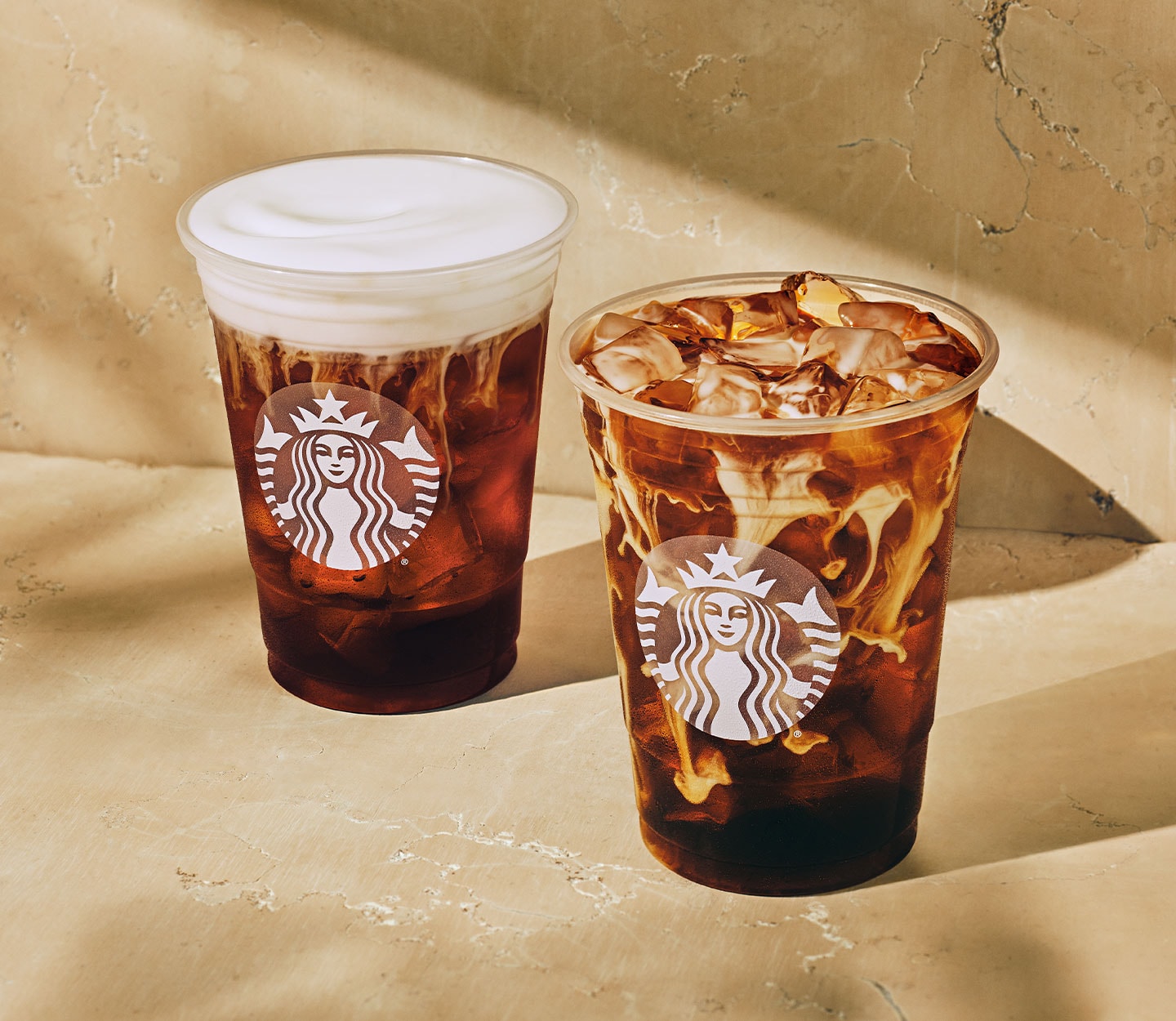Two cold coffee drinks in clear cold cups, one with a thick white foam topping, the other with creamy marbling.