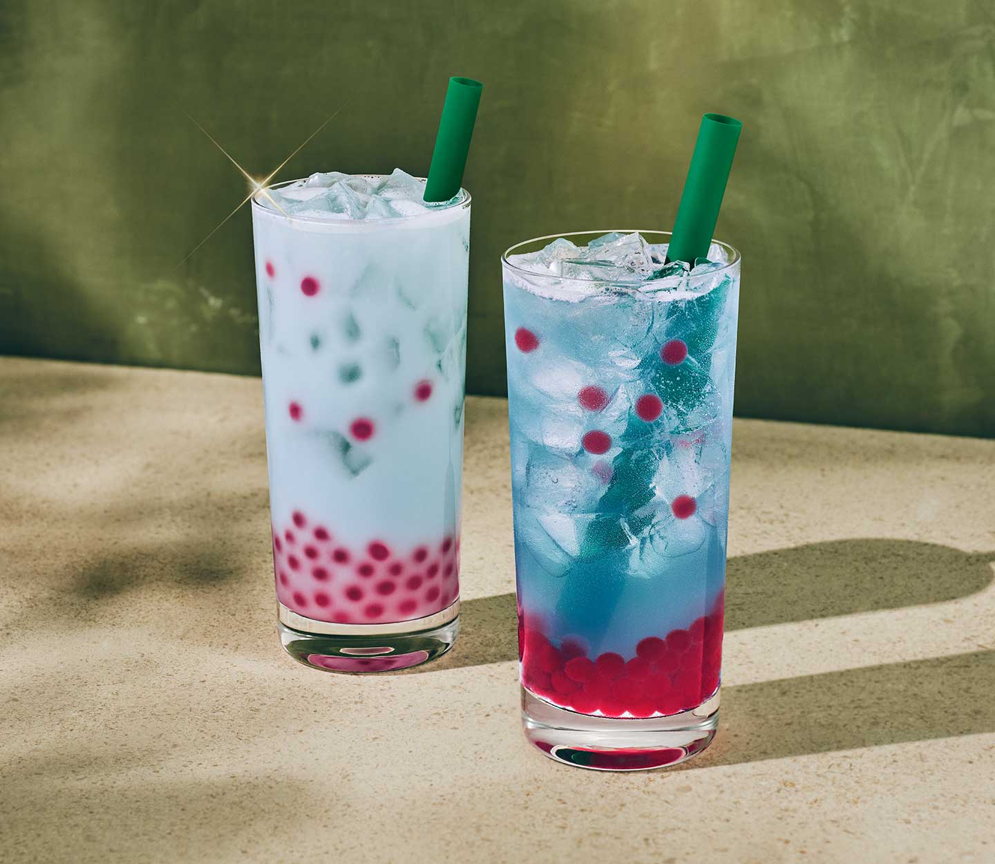 An iced pale blue drink next to an iced light blue drink, both with a bottom layer of red pearls, in tall glasses with green straws.