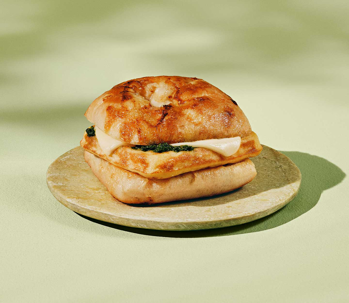 A toasted bun sandwich with an egg frittata, green pesto spread and slice of cheese in the middle, sitting on a beige plate.
