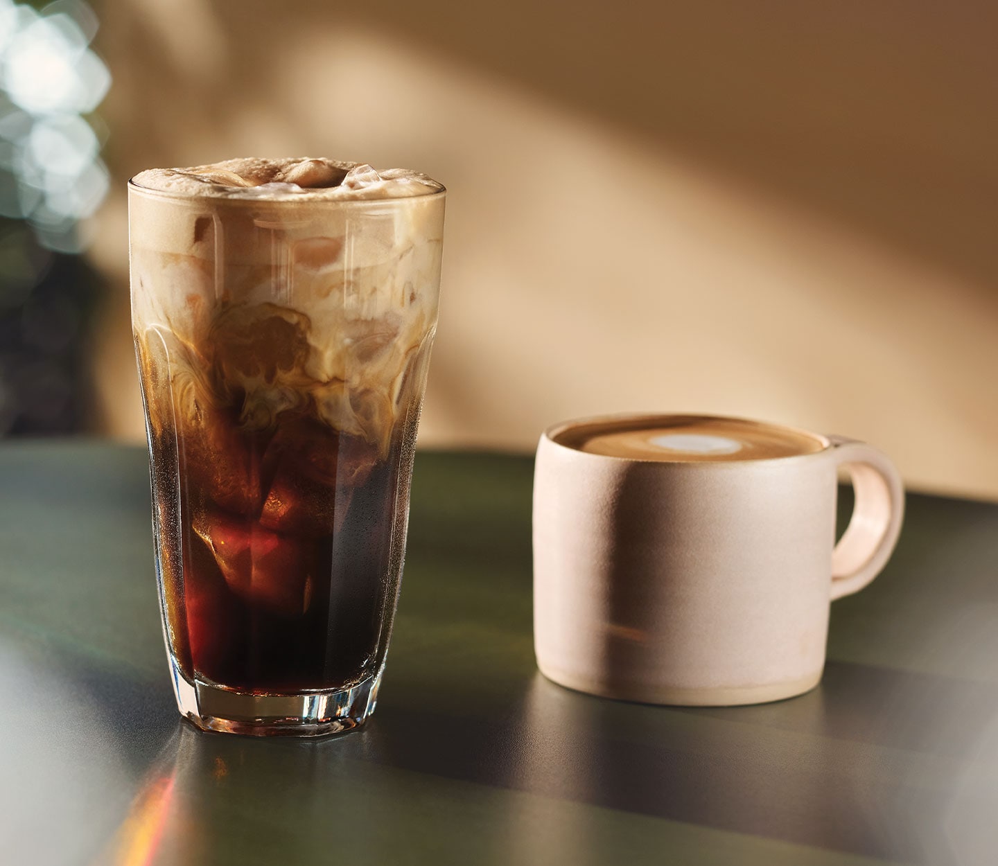 An iced coffee drink in a glass cup with swirls of milk throughout and a white ceramic mug holding a frothy coffee drink sit on a table.
