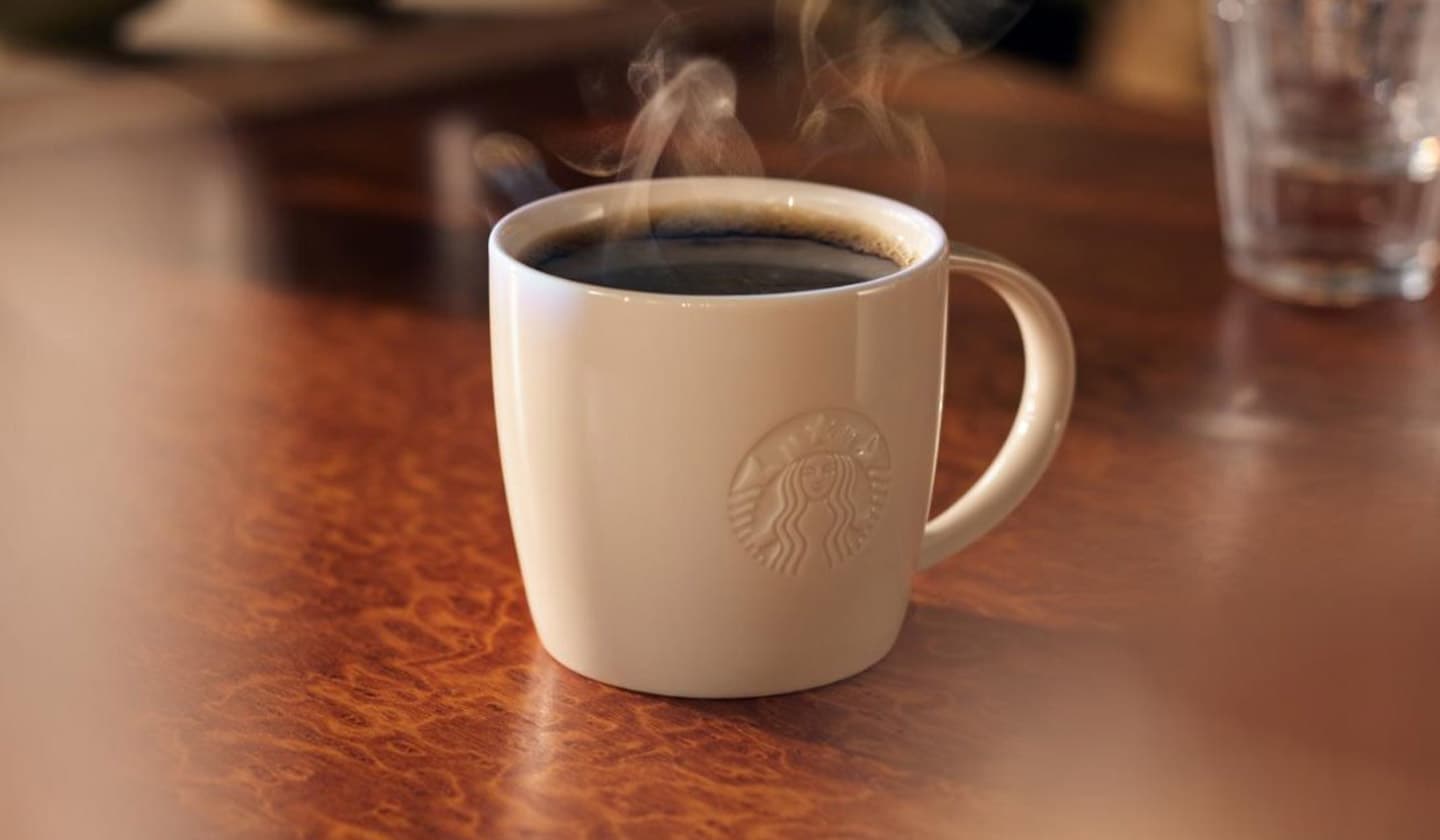 Hot coffee in a white porcelain mug with a debossed Starbucks logo is placed on a dark wood counter with glassware in the background.