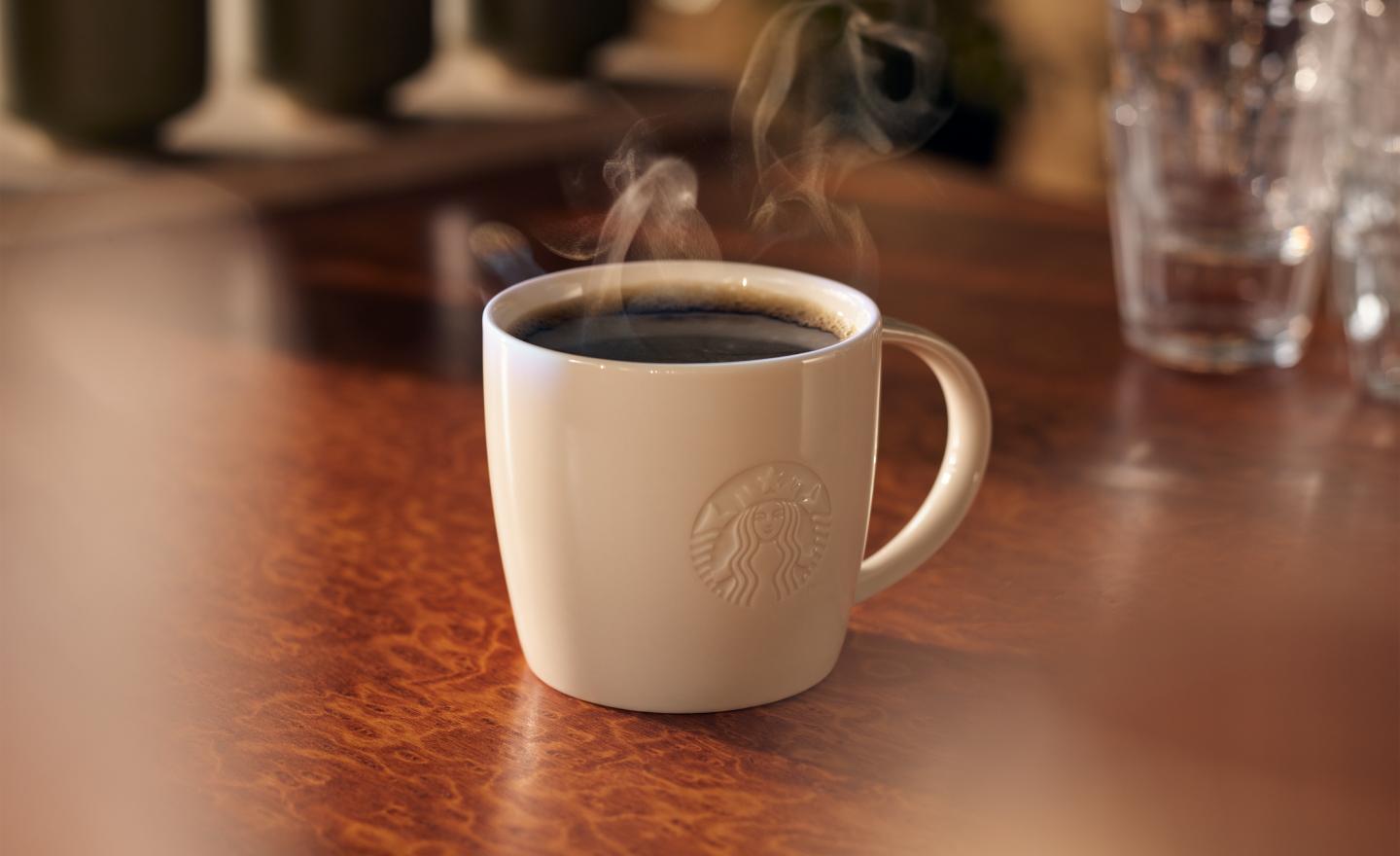 Hot coffee in a white porcelain mug with a debossed Starbucks logo is placed on a dark wood counter with glassware in the background.