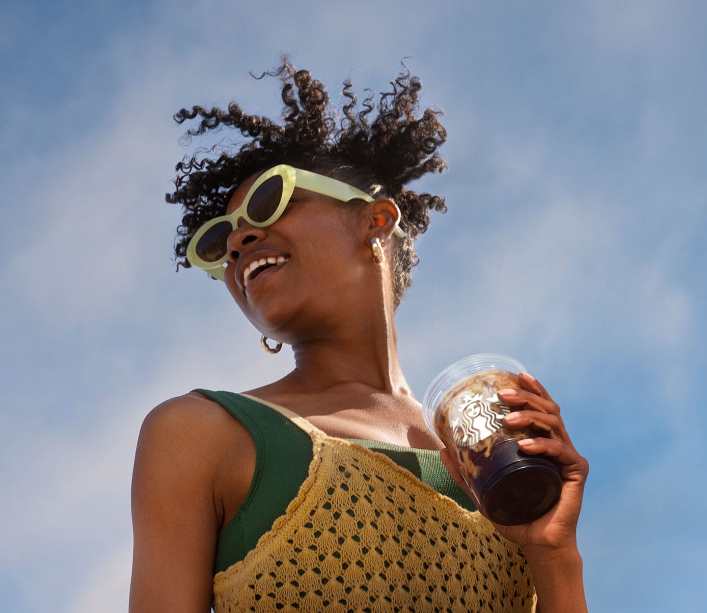 A person is outside smiling under a blue sky. She is wearing a summer tank top and sunglasses and is holding a cold coffee drink in a clear cup.