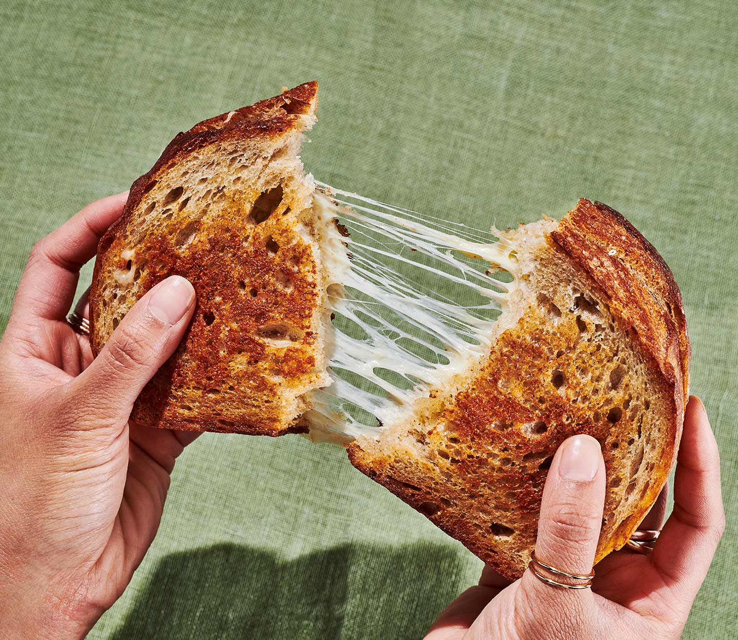Two hands pulling apart a toasted sandwich still connected by stretched strands of creamy melted cheese.