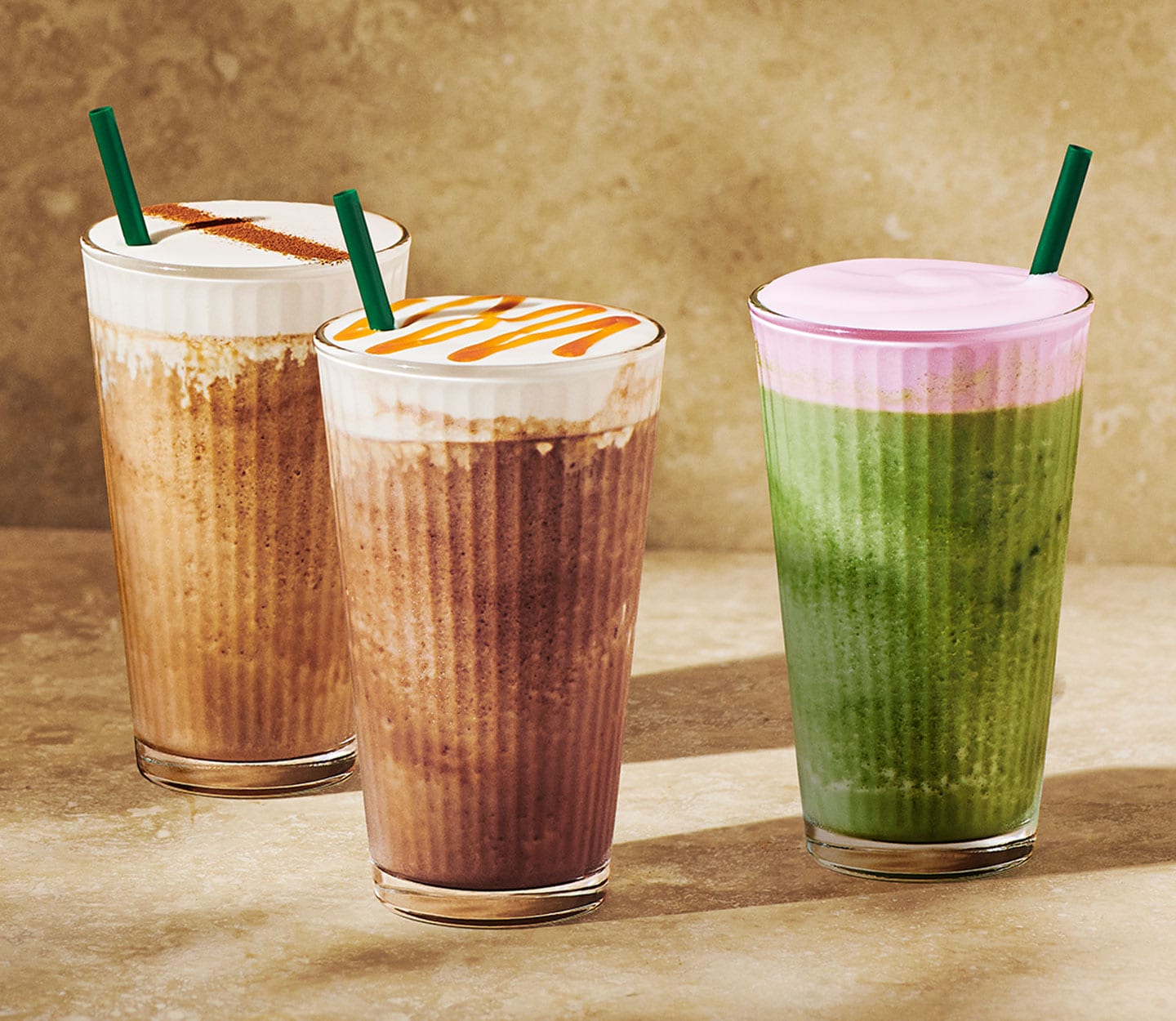 Three cold blended beverages in tall glasses with green straws. Two are coffee drinks with creamy foam toppings and drizzle. The third is a green tea drink with pink foam topping.