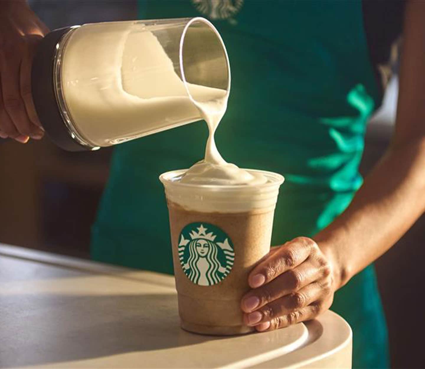 A Starbucks® barista in a green apron pours a foamy topping onto a cold blended coffee drink in a clear cup on a counter.