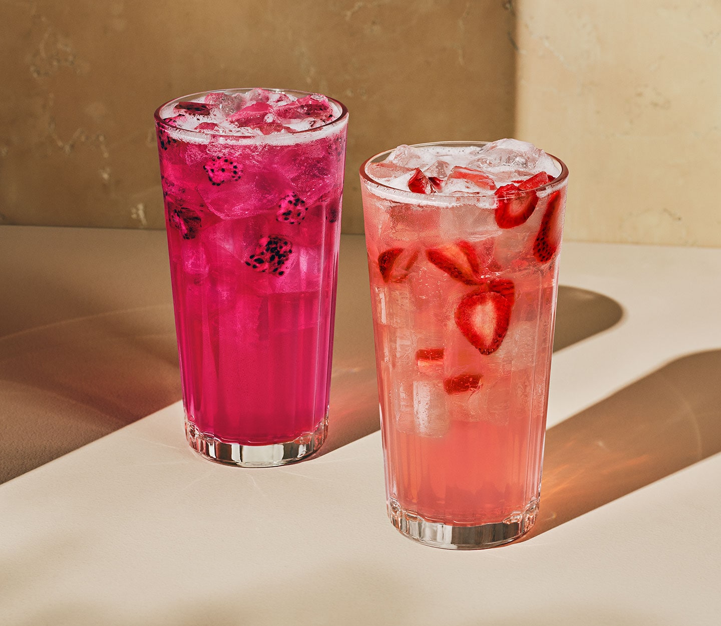 Two iced drinks in tall glasses: one dark pink with floating dragonfruit pieces, the other light pink with floating strawberry pieces.