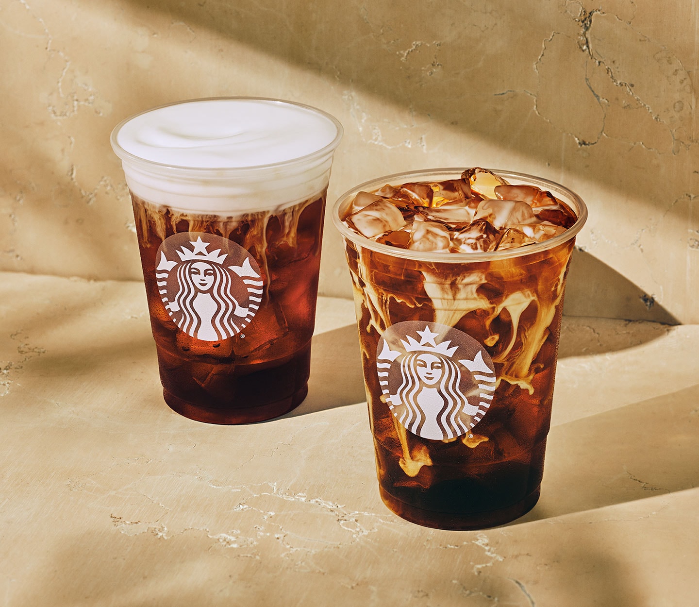 Two cold coffee drinks in clear cold cups, one with a thick white foam topping, the other with creamy marbling.