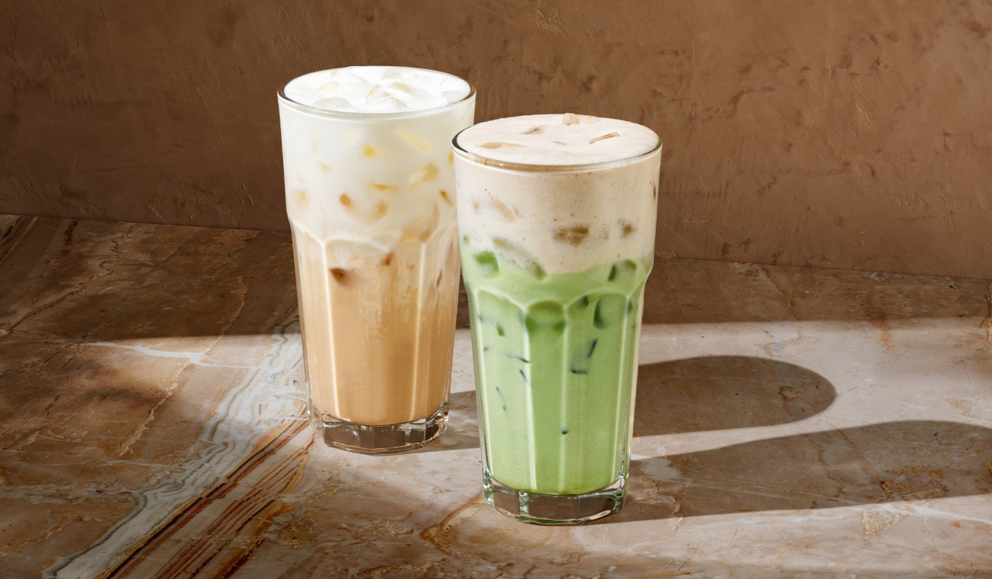 We see two iced drinks – the one on the left is a refreshing iced vanilla latte beige in colour, next to a milky-green iced banana matcha. Both are topped with a creamy protein foam against a brown background reflecting warm sunlight and the drinks’ shadows.