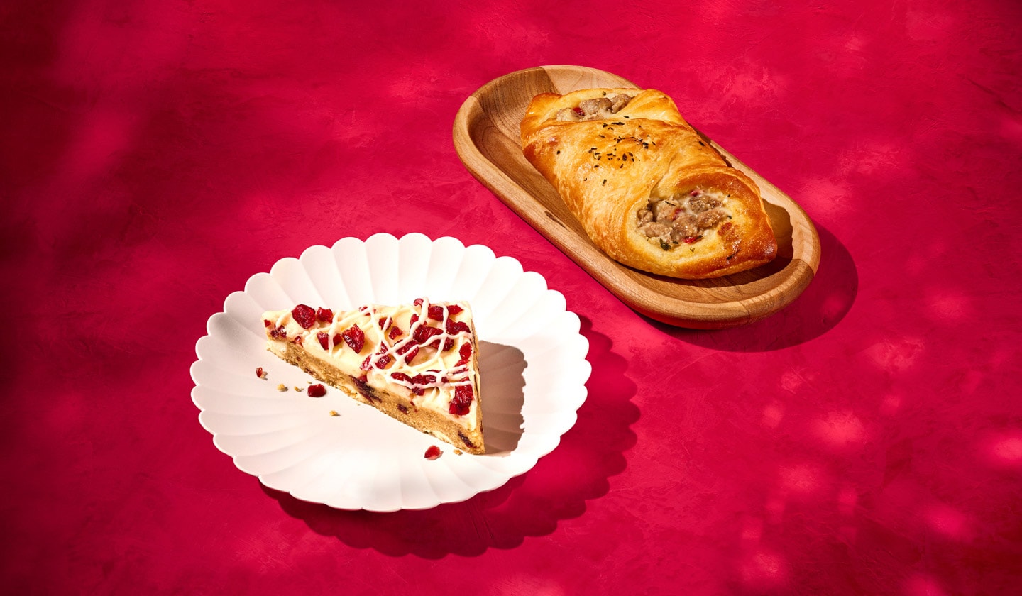 A cranberry blondie covered in a white chocolate drizzle sitting on a scalloped white dish next to a savoury oven-baked turkey danish on a wooden plate on a red velvet textured background.