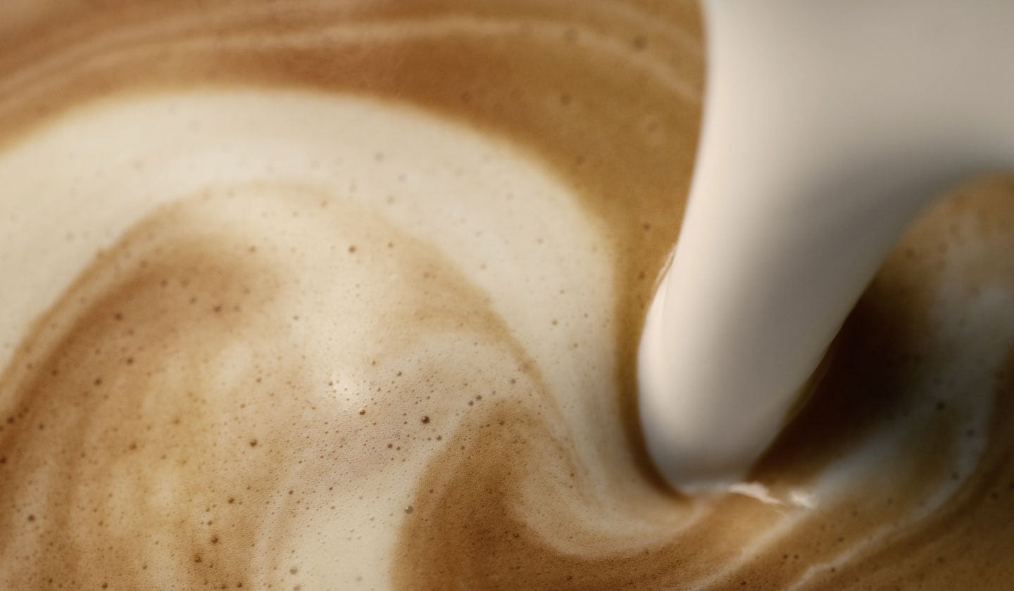 A close-up, bird’s eye view of creamy protein milk being poured into a creamy latte. There is a blend of golden espresso tones with milk cascading together.