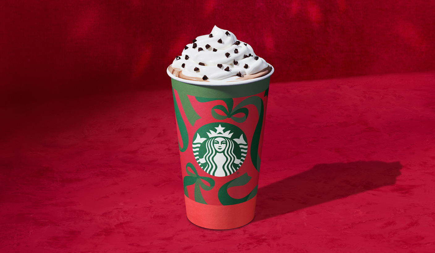 A holiday cup filled with a hot espresso drink topped with whipped cream and chocolate curls on a red background.