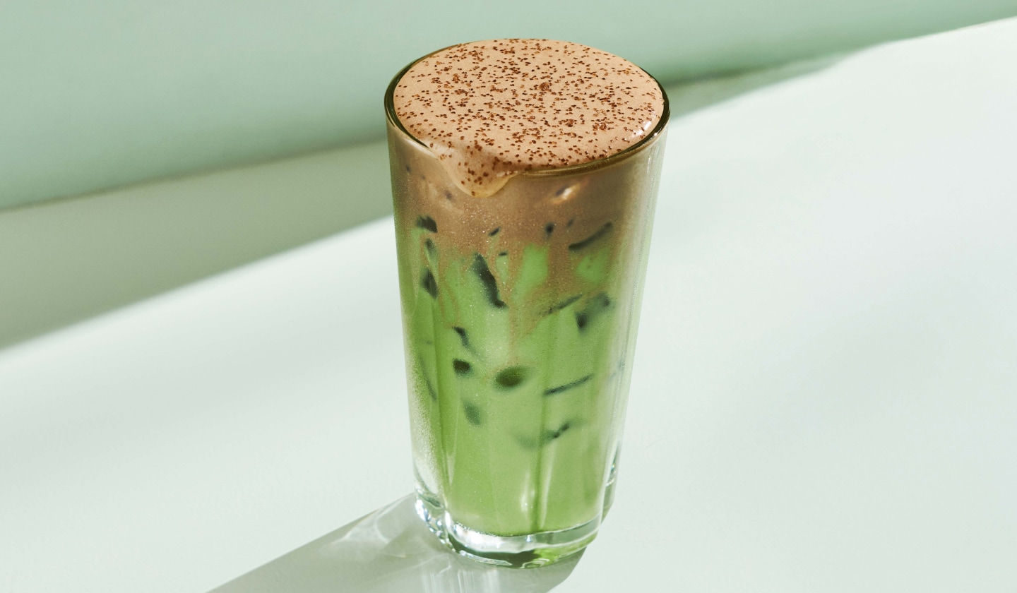 A tall glass of a vibrant green iced matcha latte, topped with chocolatey brown cold foam, dusted with brown buttery sprinkles. It is sitting against a minty green backdrop with bright reflections and soft shadows.