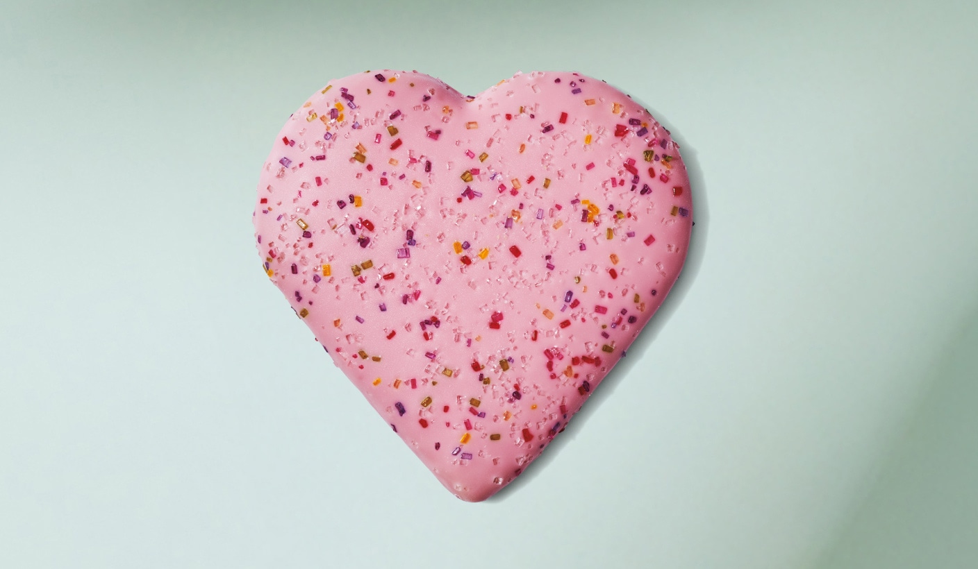 A large, tasty-looking heart shaped cookie with thick pink icing and decorated with colourful sprinkles on a minty green background.