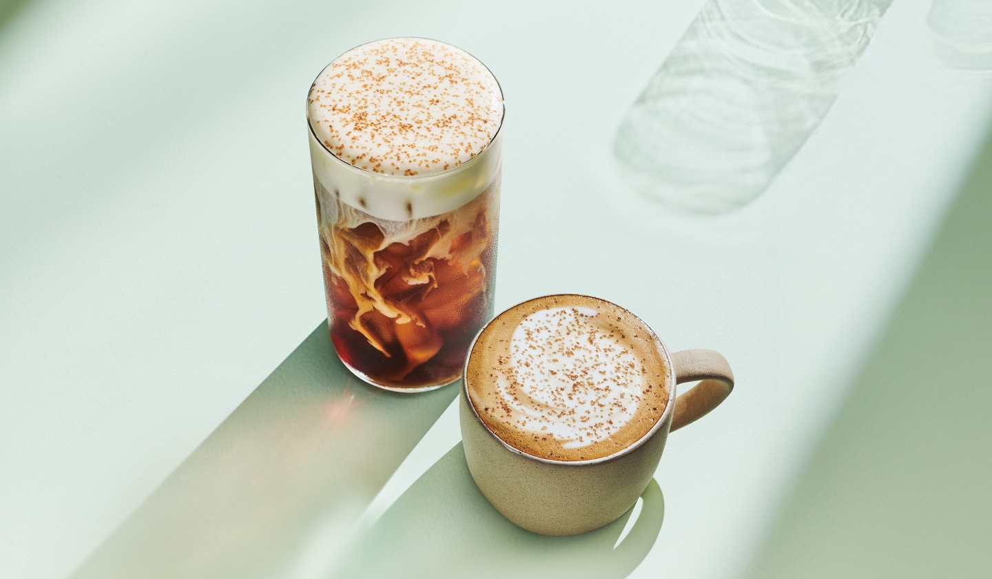 A marbled iced cold brew topped with fluffy pistachio cream cold foam and golden sprinkles sits beside a warm, handcrafted latte with swirls of golden espresso and creamy milk.