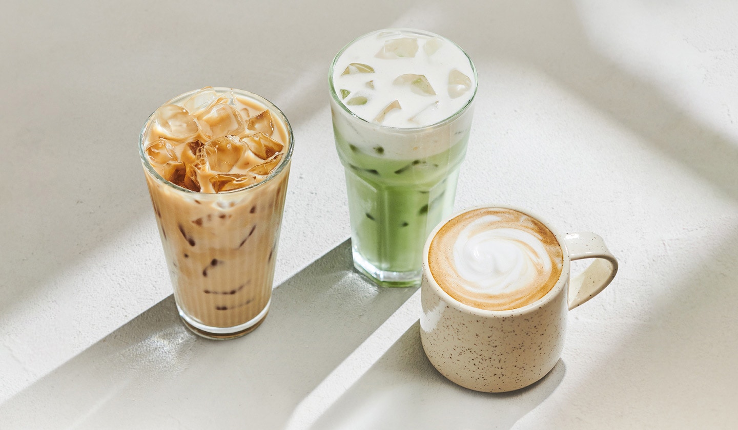 A craveable iced coffee sits on the left, a bright green iced matcha with fluffy protein cold foam stands in the center, and a latte in a beige speckled mug is on the right, all set against a white backdrop with bright reflections and soft shadows.