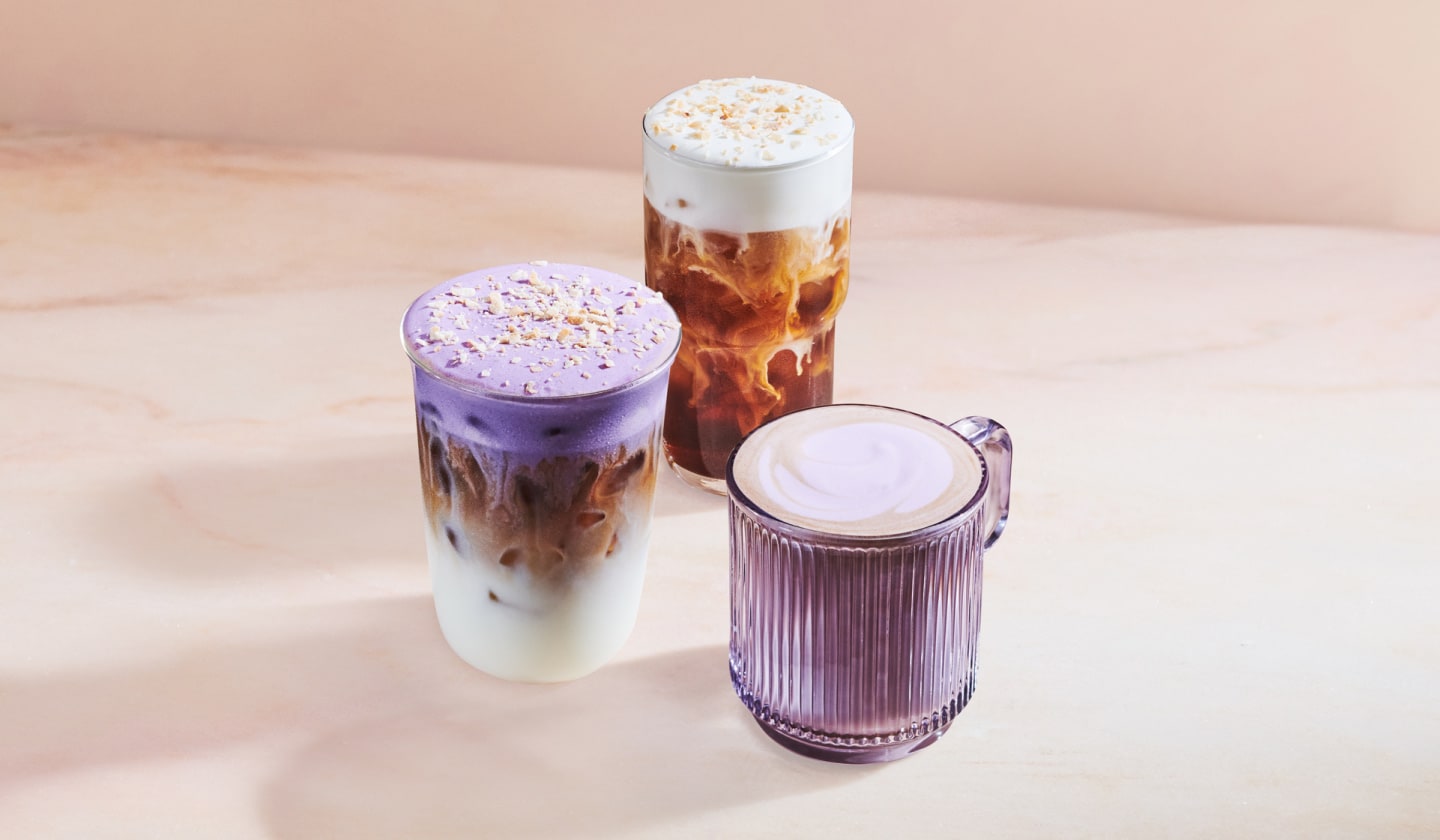 Three beverages are layered on a soft neutral background. Left we see an iced beverage with steamed milk and espresso topped with purple foam & coconut flakes. Behind is an iced cold brew topped with cold foam, with a hot latte to the right
