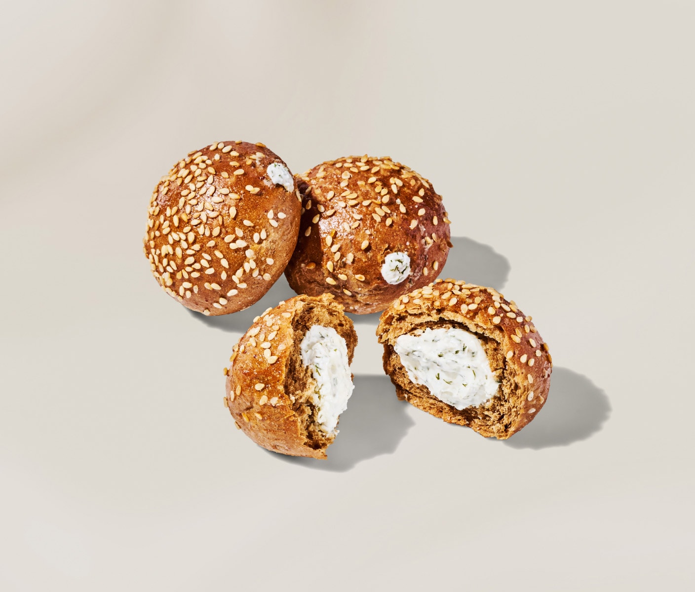 Three mini rye pumpernickel bagel rounds filled with dill cream cheese and topped with white sesame seeds on a neutral background.