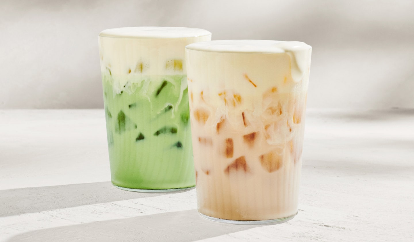 A beige iced chai latte & green iced matcha drink both topped with a creamy yellow mango-flavoured cold foam on top.