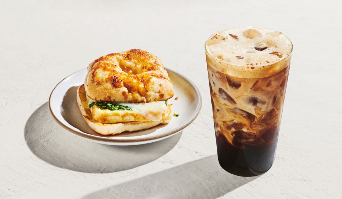 A cheese-topped breakfast sandwich sitting next to a beautifully marbled iced espresso beverage on a sun-filled white background.