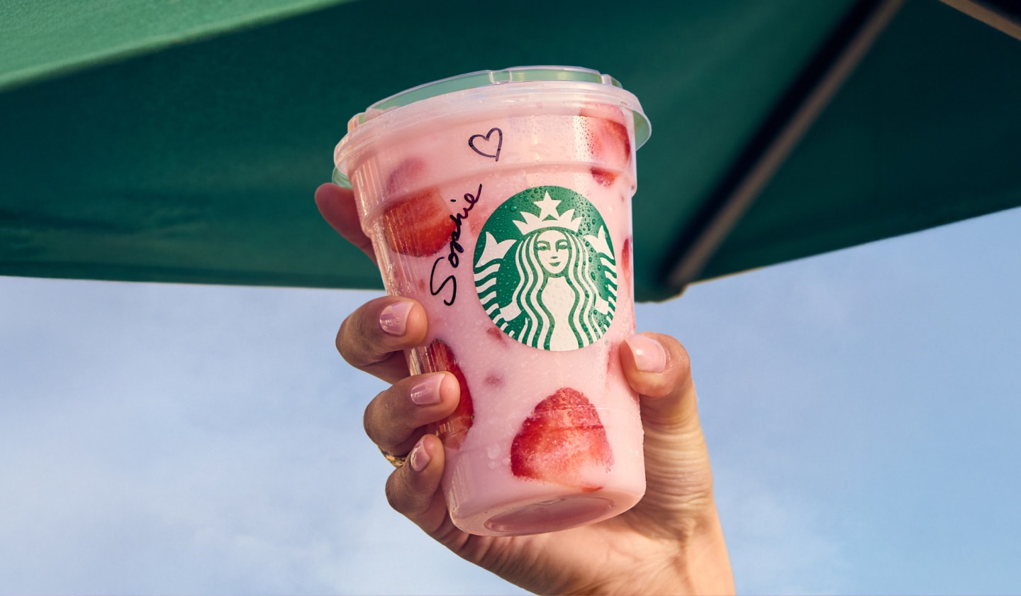 A hand holding up a refreshing pink iced beverage with dried strawberry pieces. There is a green umbrella and blue sky in the background.