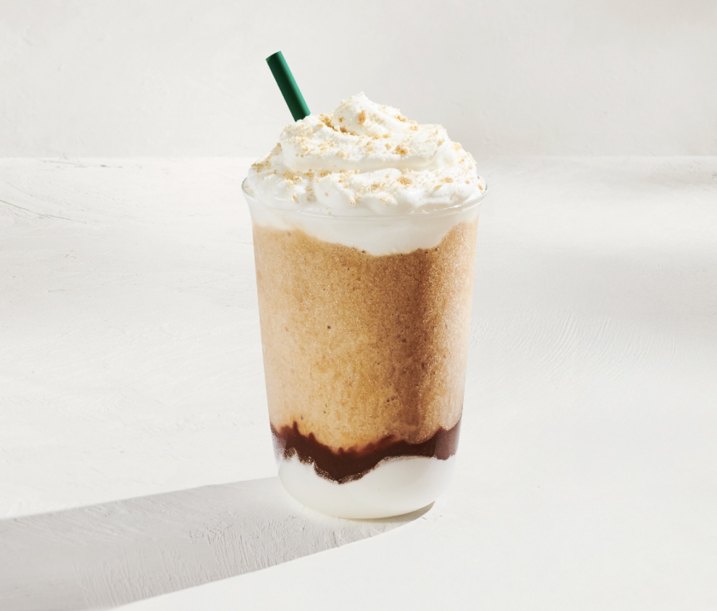 An iced blended coffee with layers of whipped cream, chocolate and blended coffee topped with whipped cream and graham cracker topping with a green straw.