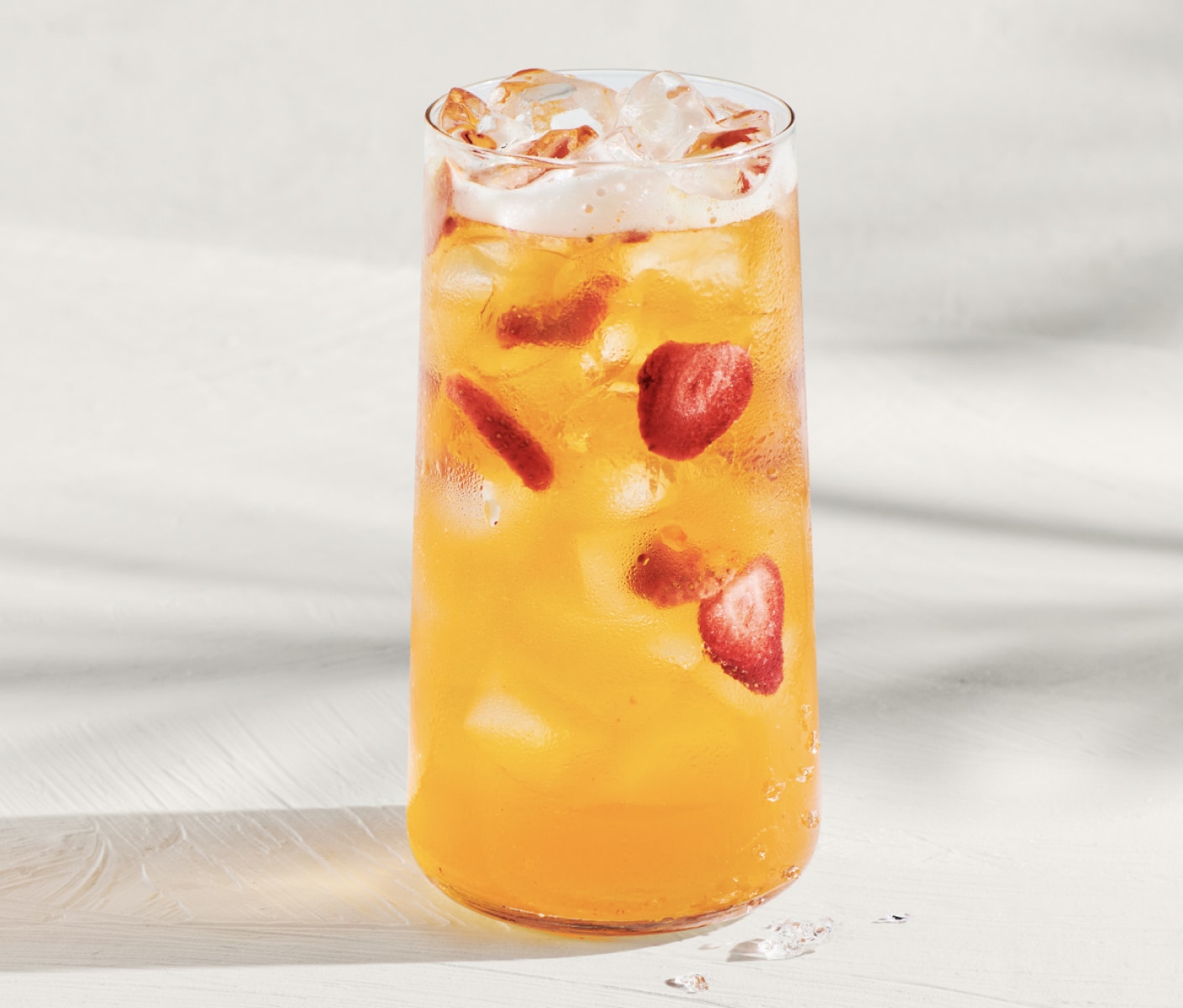 A yellow mango flavoured iced beverage with strawberry pieces and refreshing ice cubes on a sunny white background.