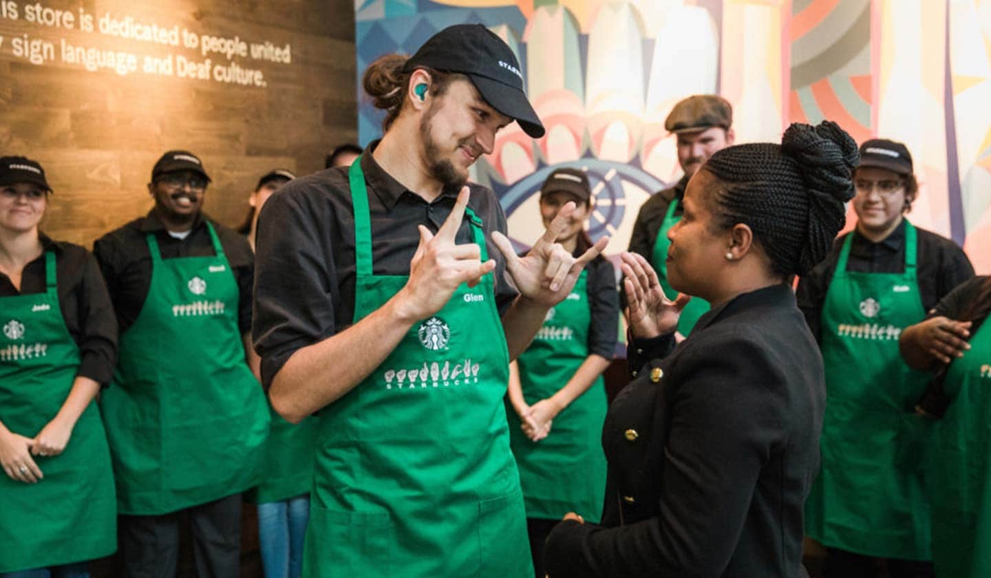 Photo of a man and woman using sign language with each other. They are wearing Starbucks green aprons with sign language signs on them and are surrounded by fellow employees.