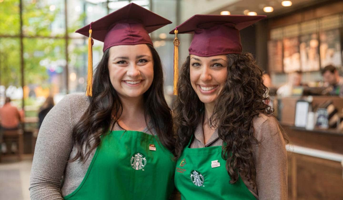 Two women in a Starbucks store smile for the camera. They are dressed in green aprons and wear burgundy graduation caps.